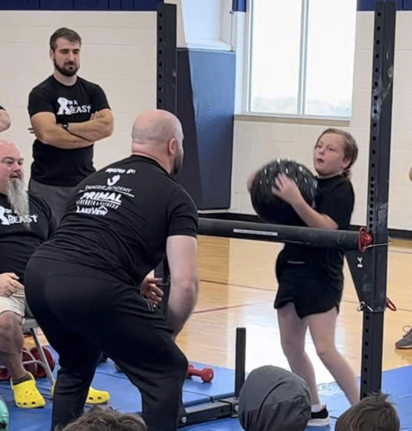 Kayla, a student at Imagine Academy and participant in Be a Beast so Families Can Feast, loads a 30 lb medicine ball over a bar, while a Strongman waits to return it back to her. 